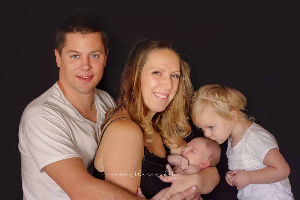 Family holds newborn daughter while older sister kisses her on the forehead